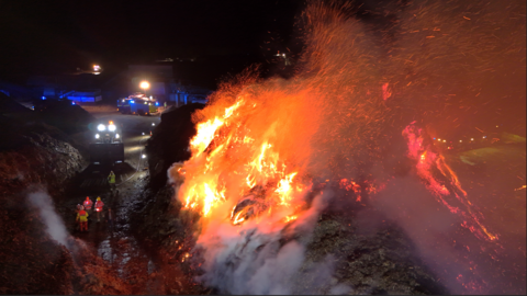 Two large piles of bark are shown with four firefighters standing between them. the left pile is smouldering but the right is fully ablaze but rest of the site is mostly in darkness other the lights of some vehicles.
