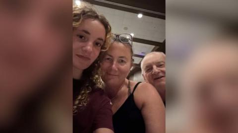 A young woman smiles next to a woman and a man. They all look at the camera.