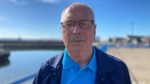 Michael Hughes stands in front of Carrickfergus marina. He is balding and wears glasses. He has a blue polo shirt on and a navy jacket over it. It's a bright sunny day and the sky is blue. 