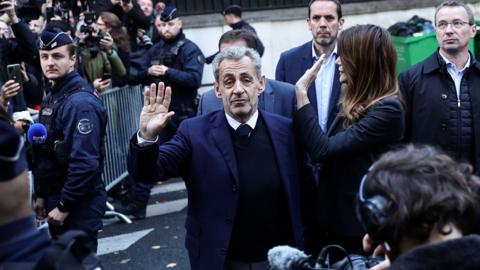 Sarkozy raises his hand as he is surrounded by members of press near a prison