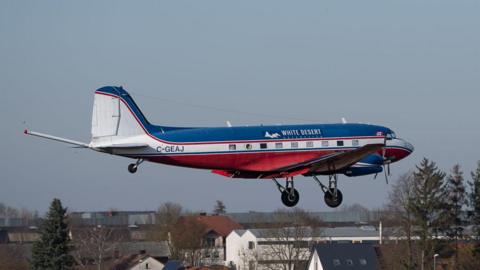 A Basler BT-67 aircraft flies low to the ground over some houses. The 1940s-looking plane is white blue and red and has "White Desert" written on the side. It has two propellers and its landing gear is down.