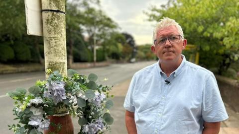 Terry Cunningham, with white hair and black-framed glasses, wears a pale blue shirt. He is standing by a lilac wreath tied to a lamppost at the spot where his mother was fatally struck by a van.