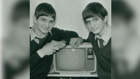 A black and white archive photograph of the Oliver twins as young men, leading on an old computer. They are wearing white collared t-shirts and ties underneath dark V-neck jumpers, smiling at the camera.