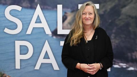 Salt Path Writer Raynor Winn with long blonde hair standing in from of a cornwall scene wit the woprds 'Salt path' written on it. She is looking at the camera smiling wearing black.