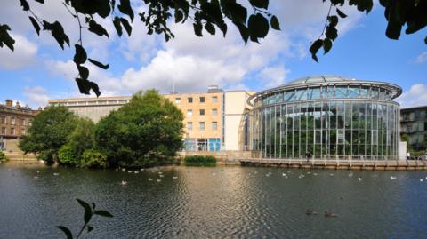 Brick building with multiple windows and  curved glass building. There are trees in front of it and a large pond.