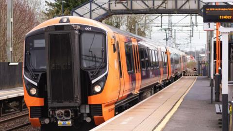 An orange and black Class 730 electric train stopped at a small railway station with a walkway over the track in the background. The platform id deserted. Trees line the other side of the track.