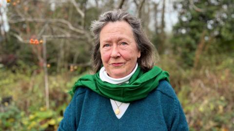 Judith Heinemann with long brown hair, looking at the camera and wearing a blue V-neck jumper over a white top. There is a green scarf around her neck. There are trees and a hedge behind her (in her own garden).