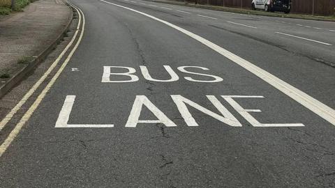 An unidentified road with white Bus Lane wording written across one lane. There are double yellow lines to one side and a thick white line on the other side  