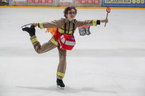 A boy with brown curly hair is skating on an ice rink, holding his back leg in the air and touching the blade of his skate. He wears the iconic brown and yellow Ghostbusters costume with a bred fire engine plushie around his neck.
