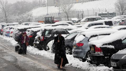 Woman walking through a car park in heavy snow carrying an umbrella