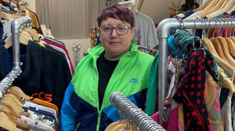 Woman with dark purple hair wearing a fluorescent green and blue jacket among rails of clothes