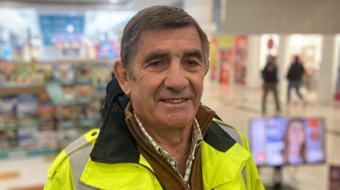 A man stands in a shopping centre wearing a high-visibility jacket with people walking in the background.