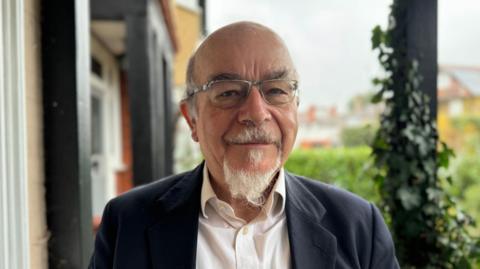 Anthony Sheppard, the chair of Claygate Parish Council's planning committee, looks straight at the camera. He is wearing a white shirt and a dark suit jacket, and has a small white beard.