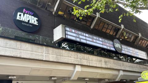 A picture of the HMV Empire Coventry, taken from street level, with hoardings advertising gigs from The Specials, Fatboy Slim and Simon Brook. A first-floor terrace has glass around it, and a promontory overhangs the second floor. A round black sign on the building says HMV Empire Coventry.