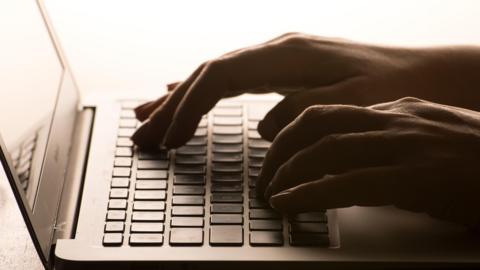 A picture of someone typing at a computer. The hands can be seen typing with the keyboard and part of computer screen visible.