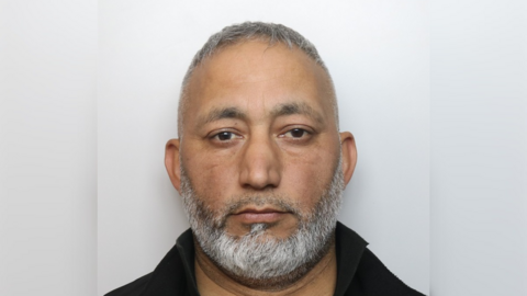 A mugshot of Muhammad Arshad. He has brown eyes and a white and grey beard. He is wearing a black top with a collar. 