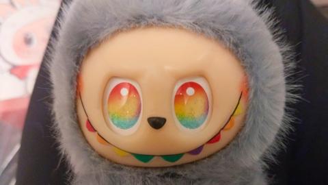 A close up of a fake Labubu doll with grey fur, and rainbow eyes and smile.