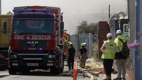 A red appliance with Fire & Rescue written across the front and blue lights flashing is parked on a road with hoses running across the street. Smoke is rising from behind a nearby fence. Several firefighters in uniform are working around the truck, and a number of other people in hi-viz jackets and white helmets are looking on.