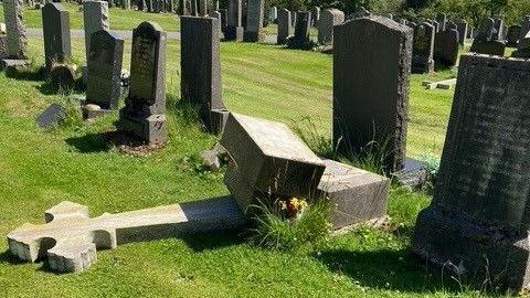 a headstone in the shape of a cross lies toppled over in front of other headstones