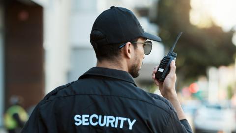 The back of a security guard who is holding a walkie talkie. The word "Security" is on the back of his black shirt and he is wearing a black cap and glasses. The background of the picture is blurred but it looks like he's on a street.