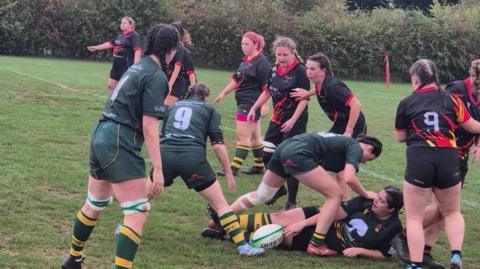 Female teams in green and yellow as well as black and red playing a rugby match.