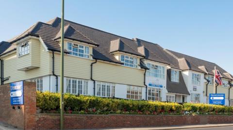 A large yellow and white two storey building surrounded by a low brick wall and flowers. Two blue signs that say 'Drumconnor' are visible, along with a Union Jack flag on a flagpole next to the building. 