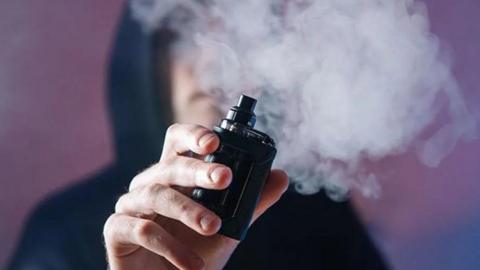 Man in hood holding a vape with plume of smoke