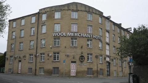A five-storey hotel on the corner of the road. A large sign says Wool Merchant Hotel