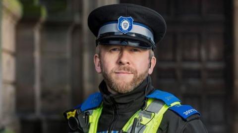 A police officer wearing a uniform and a yellow hi-vis jacket smiles at the camera. He has a brown-grey beard and short grey hair.