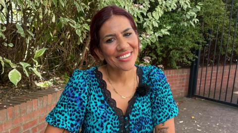 A woman with dark hair and a blue animal print dress smiles sitting in a garden.