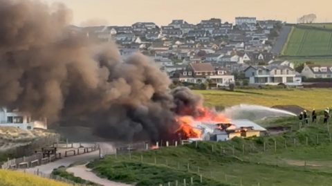 A beachside building is on fire with large plumes of black smoke coming away from it and over the beach. Fire crews are seen spraying the property with water.