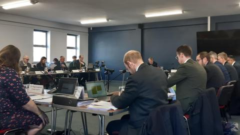 A group of people sit around tables laid out in a U shape in a room. Some of them are using laptops and have sheets of paper. 