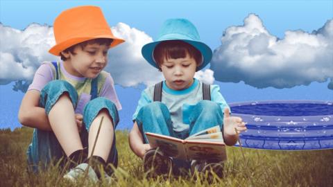 Two children read a book together on the grass, one wears an orange hat and one wears a blue hat