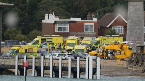Ambulances, emergency response vehicles and an air ambulance helicopter are gathered near the seafront during an emergency operation.