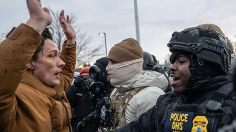 A demonstrator in a brown jacket holds their arms above their head in front of a uniformed official
