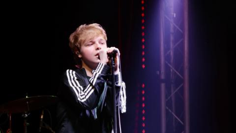 Noah performing on stage. He is wearing an Adidas jacket and sings into a microphone.