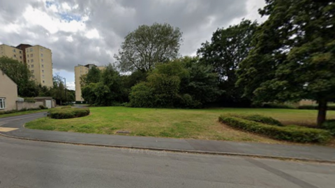 A road in the foreground, with grass, trees and tall buildings in the background.