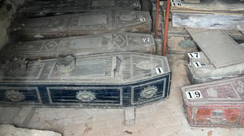 Ornate coffins in a line in a small crypt, some piled on top of others. The coffins are dusty