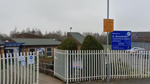 A view of a school with white gates outside it. A blue sign saying 'Welcome to St Bernadette's Catholic Primary School and Nursery' is visible, as is a yellow 'No stopping' sign which has times on it.