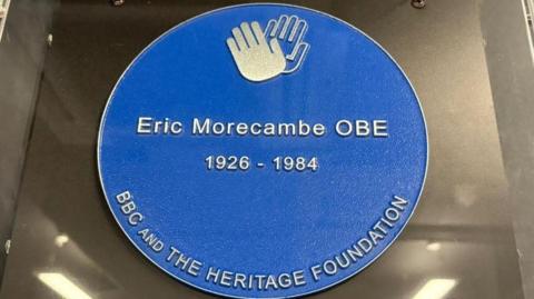 A dark blue circle plaque with two silver waving hands at the top and writing covering the rest. It reads "Eric Morecambe OBE 1926-1984" and beneath it reads "˿ and the Heritage Foundation". 