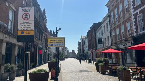 A quiet high street. There are two signs on the street, one saying "Pedestrian and Cycle Zone" with loading times underneath it and a sign next to it saying "enforcement will commence 1st June 2026". It is a sunny day with a clear blue sky. 