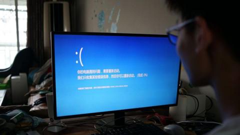 A man looks at a computer screen, which shows a blue screen with a sad face emoji. It is an error screen showing a computer has crashed, with Chinese text