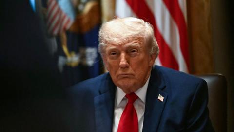 Donald Trump looks straight ahead with his eye narrowed. He is wearing a navy suit, white shirt and red tie with an American flag pin on his lapel. In the background is an American flag.