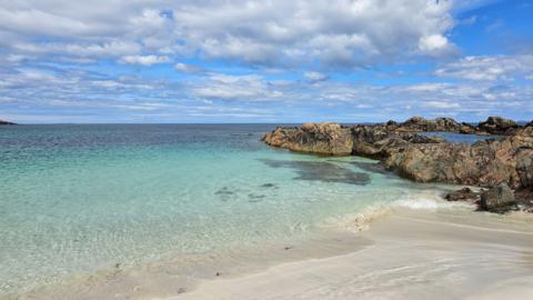 A light-colored sandy beach is in the foreground with clear, shallow turquoise water coming onto the shore. Dark-coloured rocks are to the right of the image between the sand and the water. The background displays a bright blue sky with fluffy white clouds.