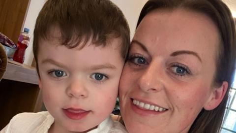 A little boy with short brown hair and his mum who has long brown hair are both smiling. They are in a room and a bottle of a Ribena drink is on a shelf behind them. There is also a window behind them.