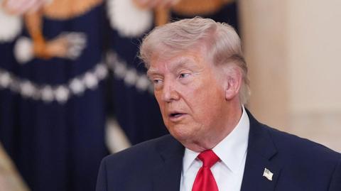 US President Donald Trump speaks while wearing a dark suit, white shirt and red tie. He stands indoors front of flag with a the US presidential seal on it.