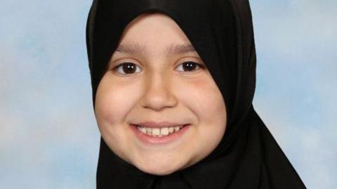 Sara Sharif smiling in what looks like a school photo. She is looking down the camera and wearing a hijab.