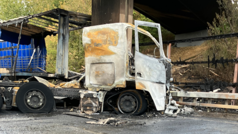 A trailer and lorry following a collision where it struck a bridge and caught alight. Some of the structure of the lorry remains in tact however the rest is charred or has perished. There is some damage to parts of the bridge and there is ash and fire remnants left on the road next to it.