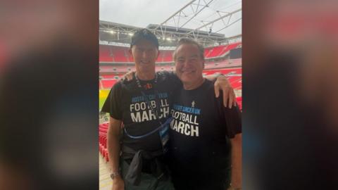 Paul Dennington and Jeff Stelling look into the camera and smile while embracing each other. They have arms round each other and are both wearing tops for Prostate Cancer UK.