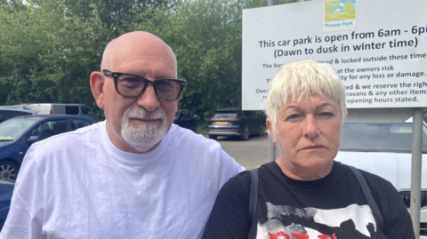 Man with bald head and grey goatee beard wearing a white t shirt and glasses is standing next to a woman who has blonde short hair and is wearing a black T shirt with a black, white and orange picture on the front. They are standing in a car park with a sign behind them which states that the car park is open from 6am til 6pm. There are large trees in the background.